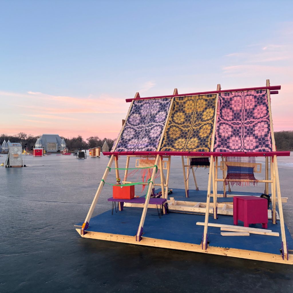 An A-frame shanty with a woven roof sits on a frozen lake against a pink and blue sky.