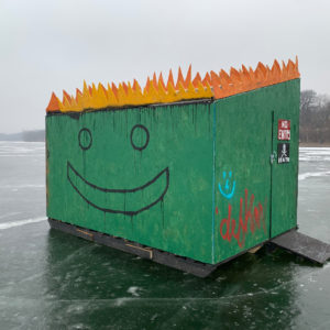 A photo of a green dumpster-shaped shanty with orange flames coming out of the top and smilley face graffiti on the side.