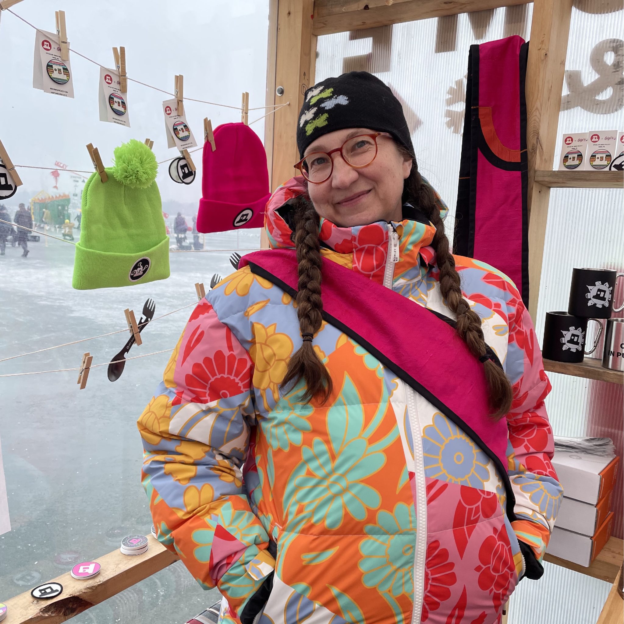 A person with two long braids wears a very colorful snowsuit and pink sash inside a shanty with merch displays.