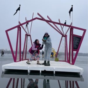 Two people perform an improv scene on a magenta and white stage flying black flags. A chalkboard lists a schedule.