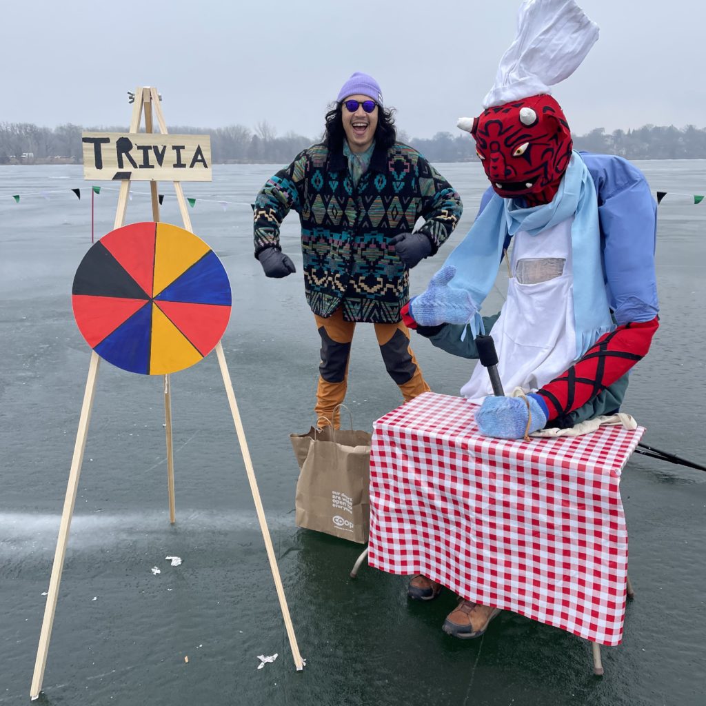 A dancing person and a giant chef puppet stand next to a colorful trivia wheel on a frozen lake.