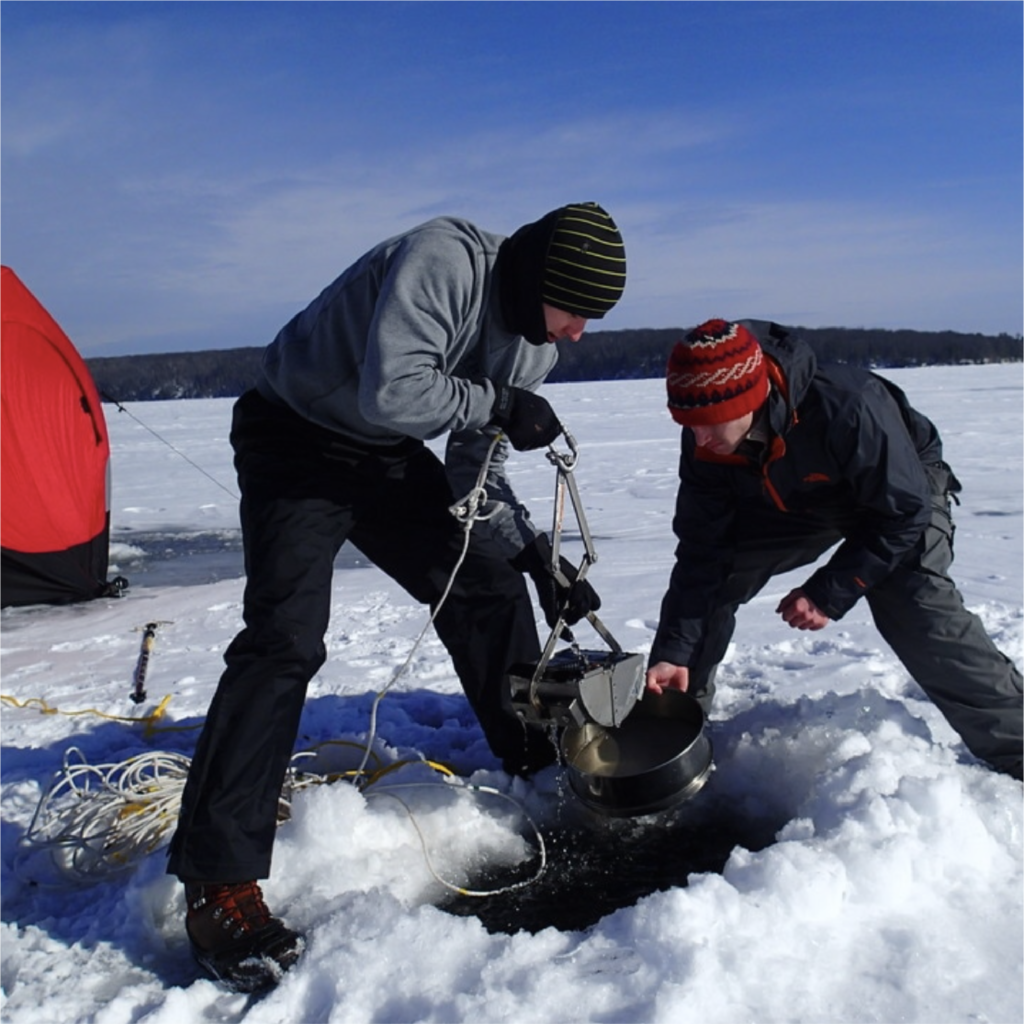 two people work together to collect water from a hole cut in the ice of a frozen lake. A red temporary ice house is in the background.