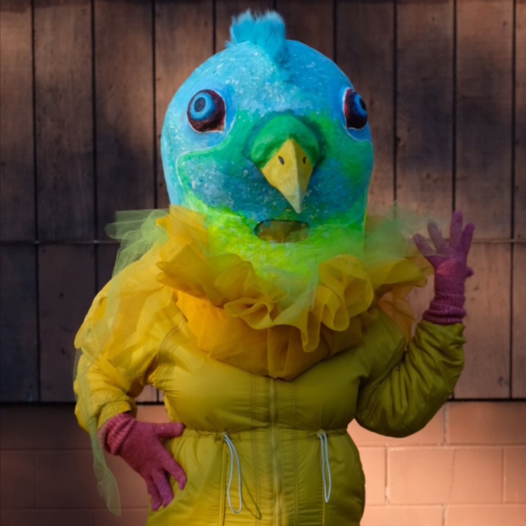 An aqua pigeon mask on a person wearing a fluffy yellow costume