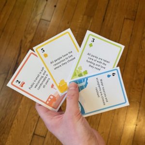 A hand holds a set of four color-coded playing cards displaying prompts about transit, housing, parks and water