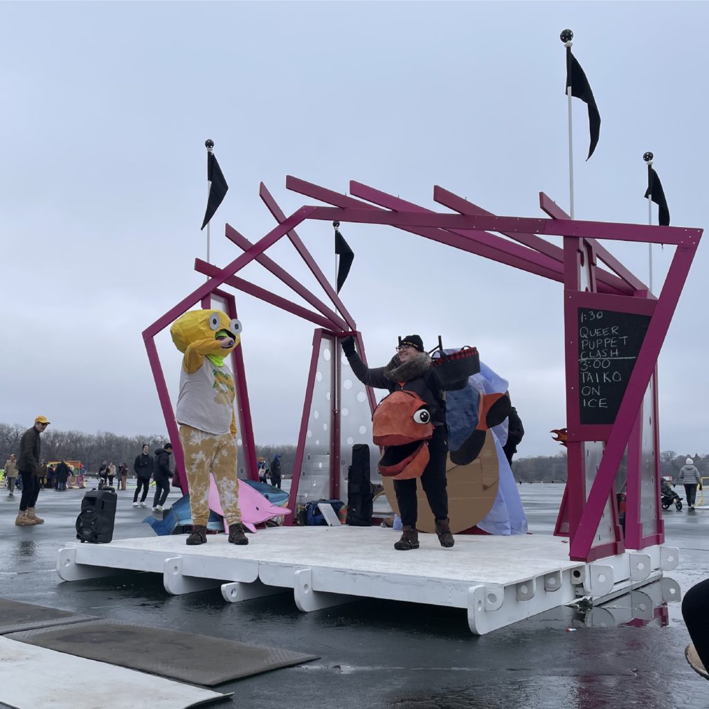 Giant puppets perform antics on an angular stage resting on a frozen lake.