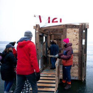 A person hands out drumsticks to people approaching a shanty constructed of a mishmash of repurposed materials