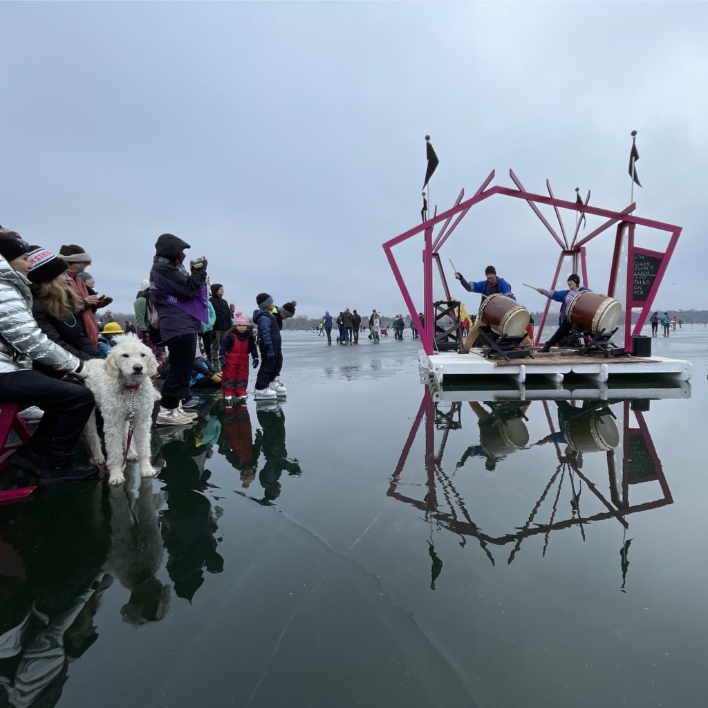 People watch a taiko drum performance on an angular magenta and black stage. The scene is reflected in the wet ice of the frozen lake they're resting on.