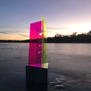 A photo of an acrylic sun dial. Light glows through the magenta and yellow panels at sunset.