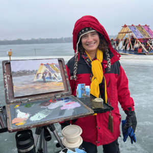 a person in a red hooded parka poses near an oil painting setup on a frozen lake. shanties are in the distance