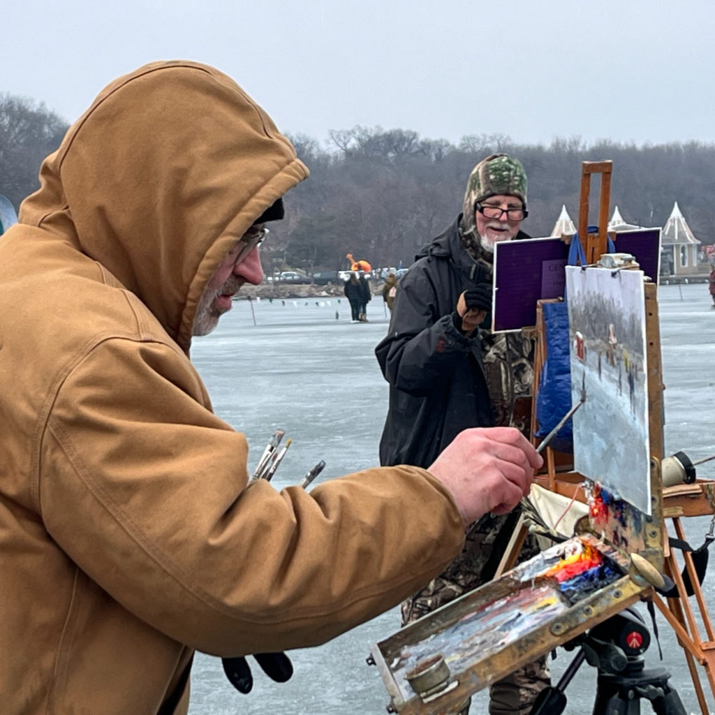 A person in a brown hooded parka paints a picture at an easel set up on a frozen lake