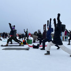 A group of people in snowsuits practice a sun salutation during a yoga session on the frozen lake