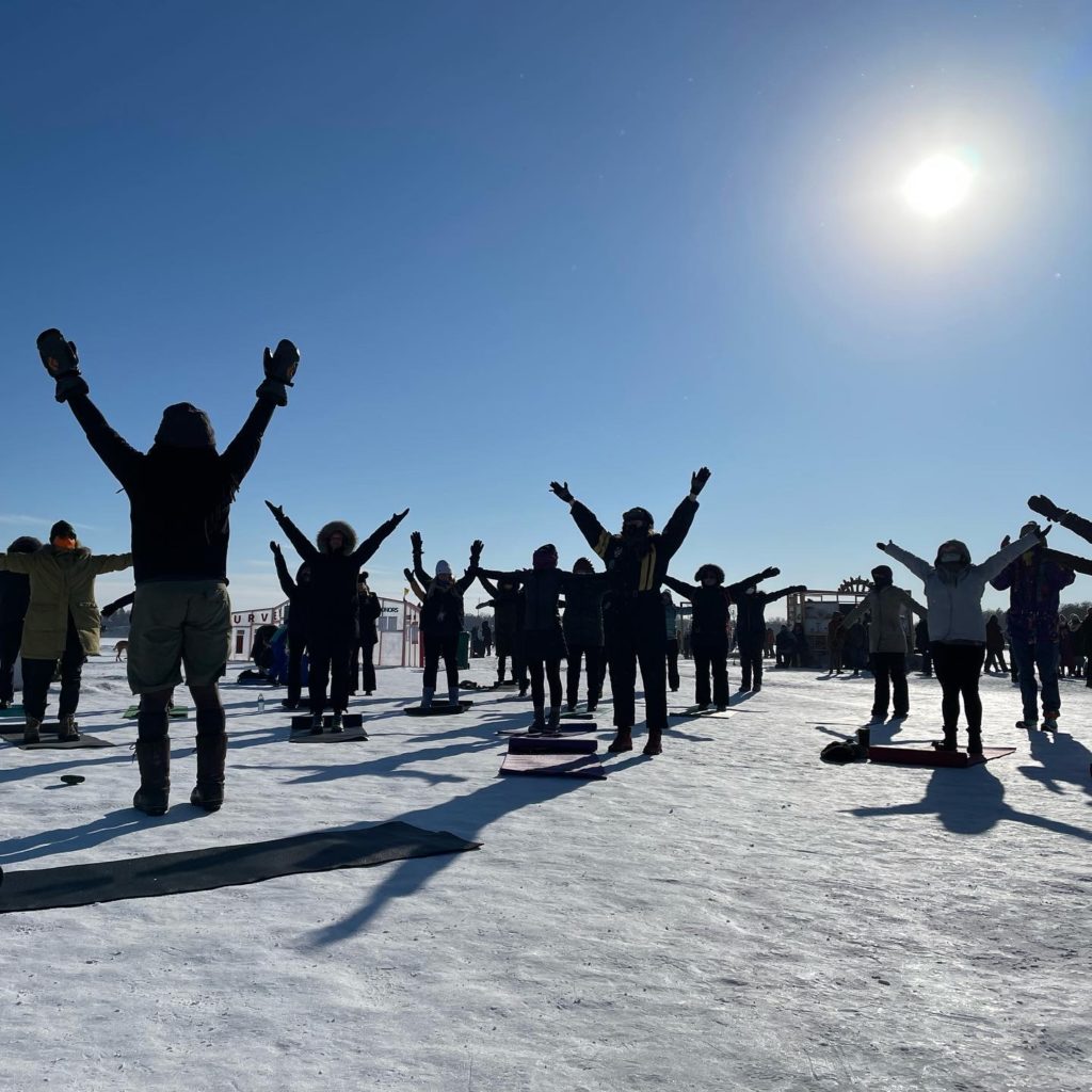 a group of people practice yoga on the frozen lake against blue skies and bright sunshine.