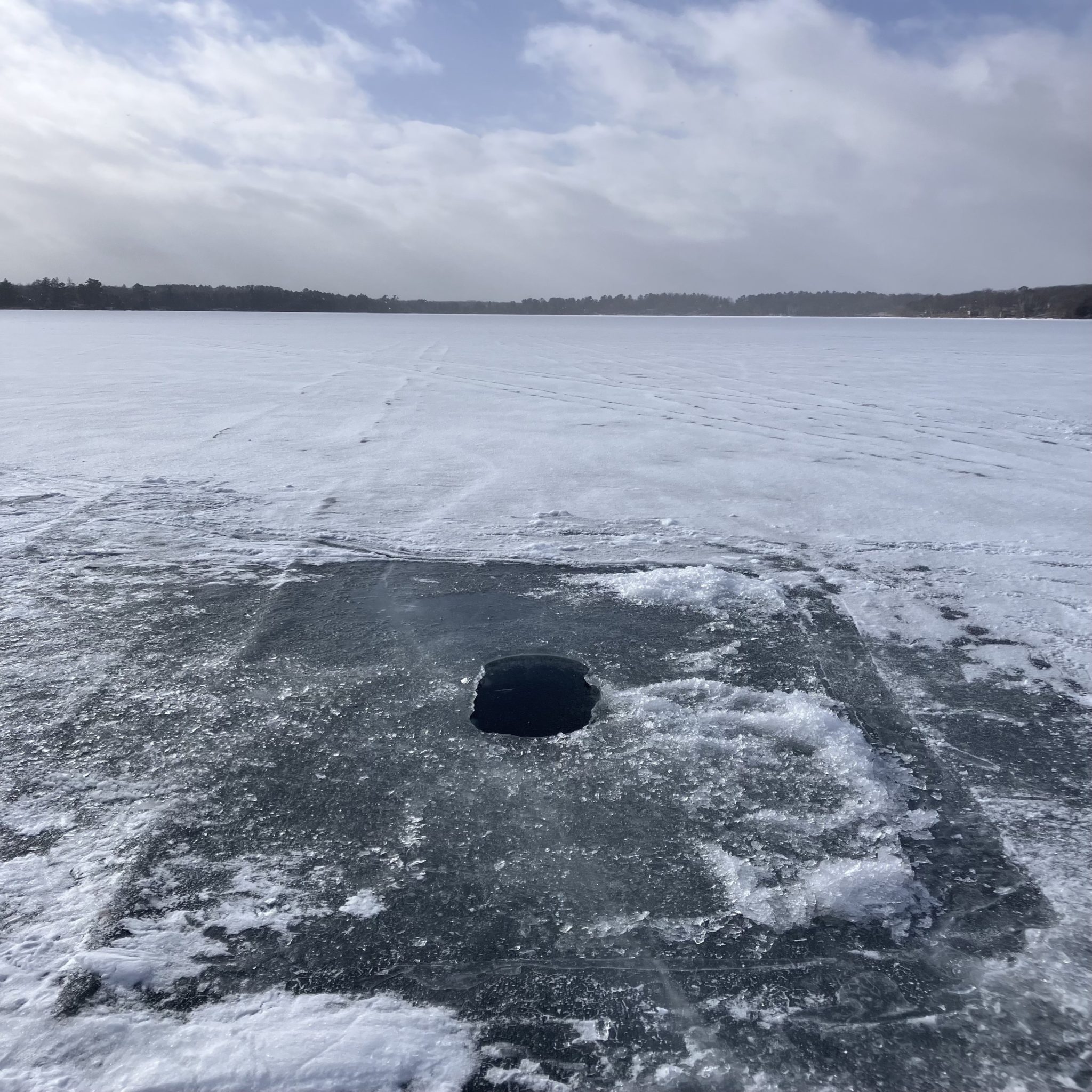 A photo of a frozen lake. Ice is shiny where a shanty had once been, with snow surrounding it. An augered hole is in the center.