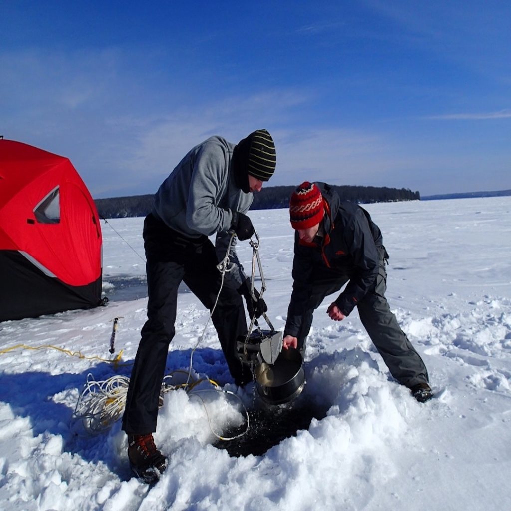 two people work together to collect water from a hole cut in the ice of a frozen lake. A red temporary ice house is in the background.