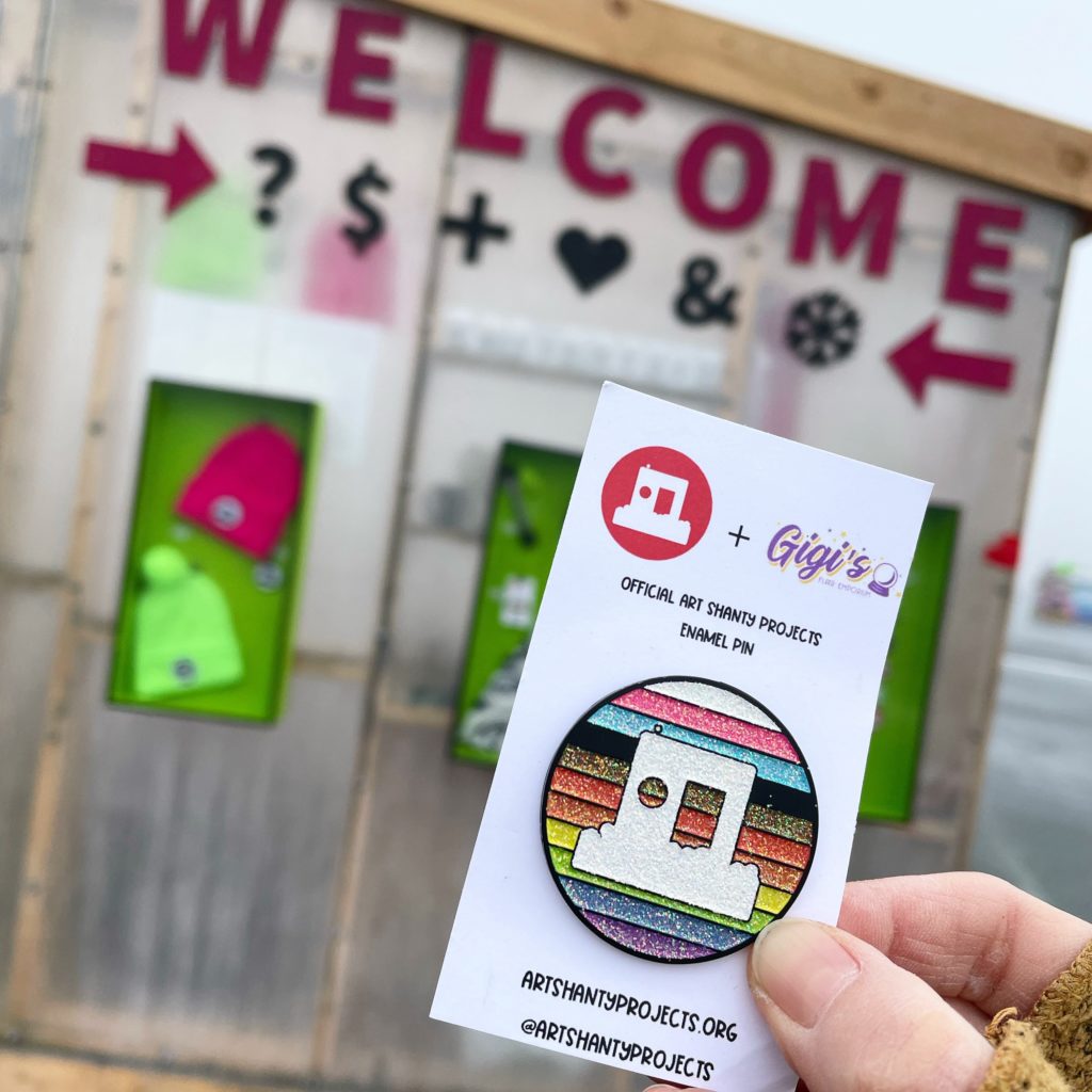 a hand holds a sparkly rainbow pin of the shanty logo. The Welcome Shanty is in the background of the photo.