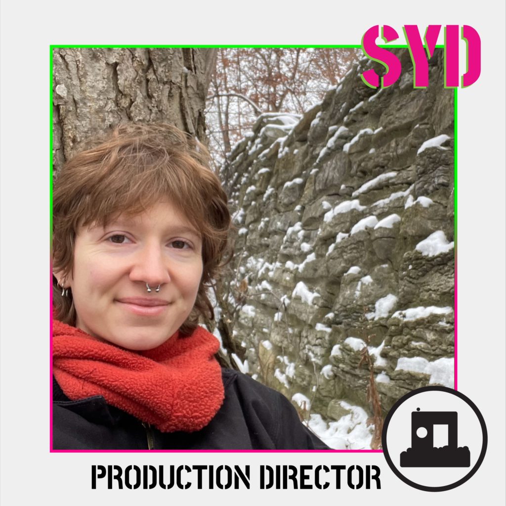 A portrait of a person with red hair standing outside in a snowy landscape. Stenciled font reads 'SYD' and "Production Director"