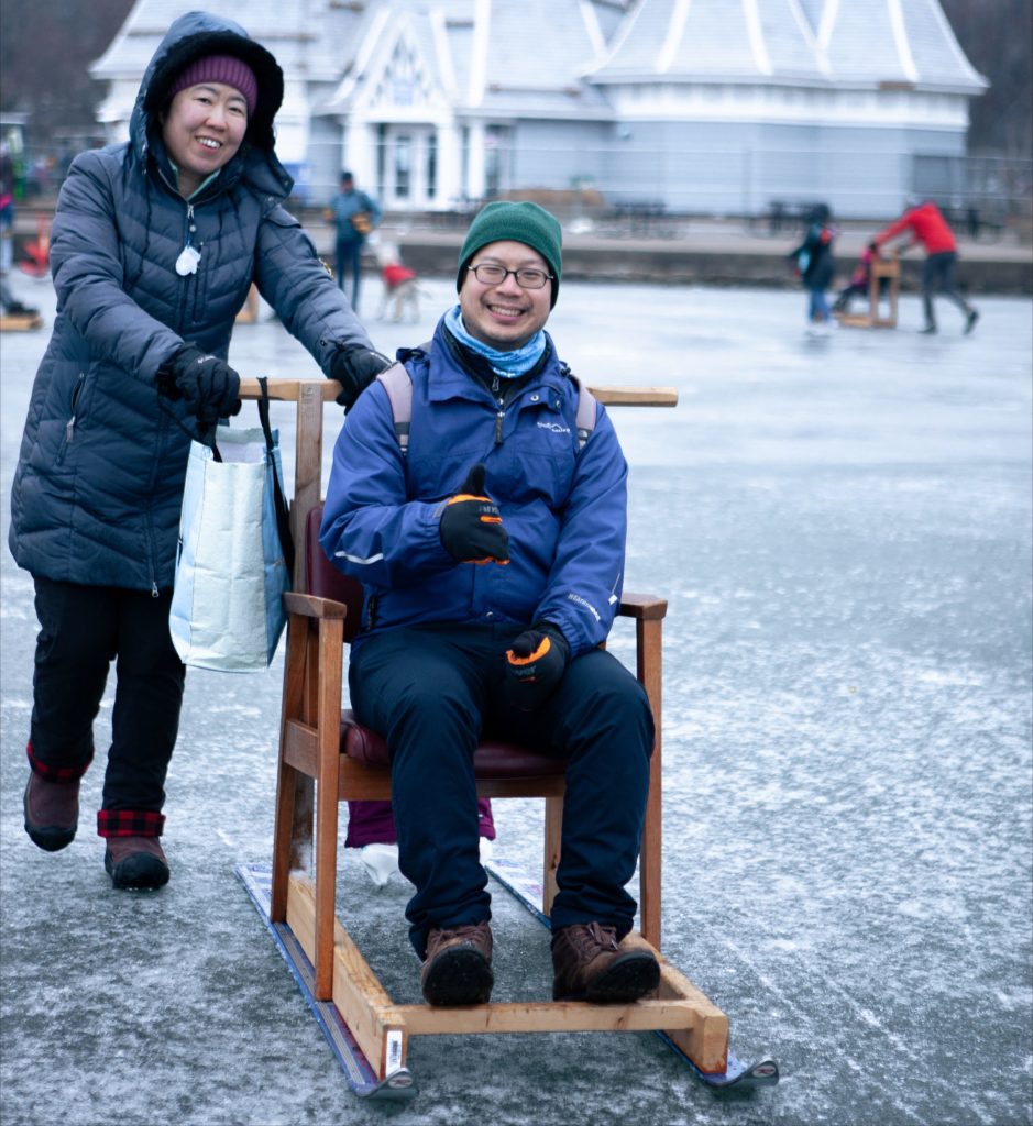 A person takes a ride on a DIY kicksled on a frozen lake. The Lake Harriet Bandshell is in the distance.