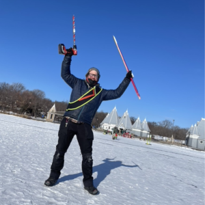 a person pauses on the ice holding up a wooden pole and a drill with an auger bit attached