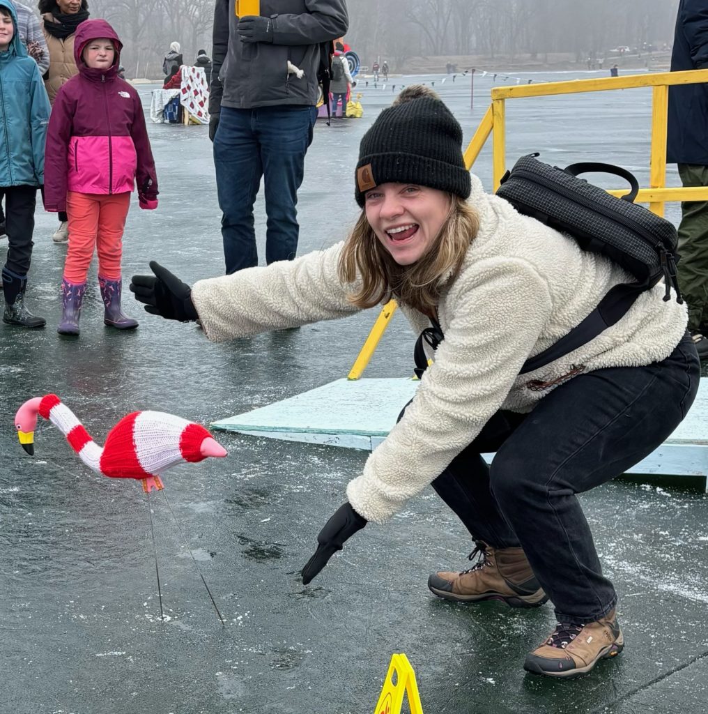 Kate Zellmer on the frozen lake excitedly gesturing at a lawn flamingo wearing a sweater.