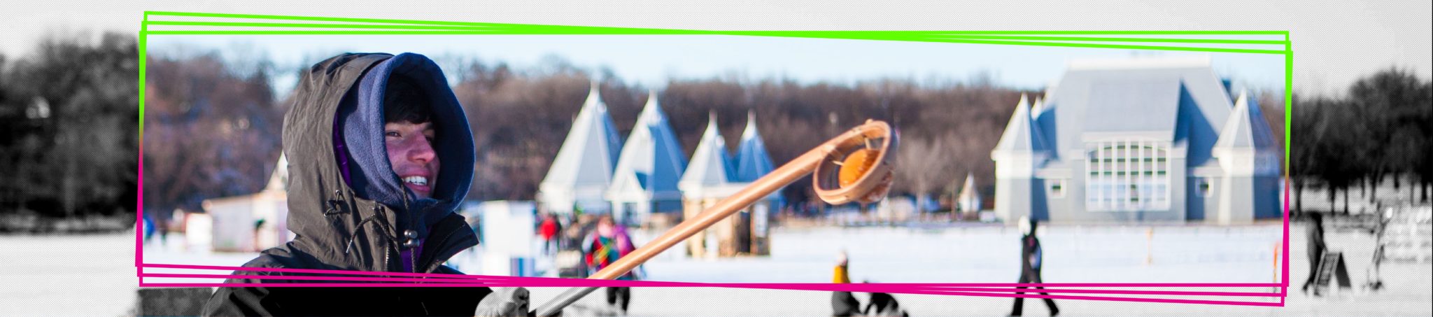 A person smiles as they raise a traditional wooden lacrosse stick on the frozen lake. The shanty village and Lake Harriet Band Shell are in the distance