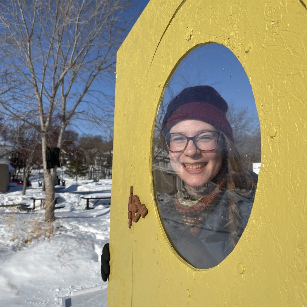 Karly is smiling while looking through a round window of a yellow shanty. Snow is in the background