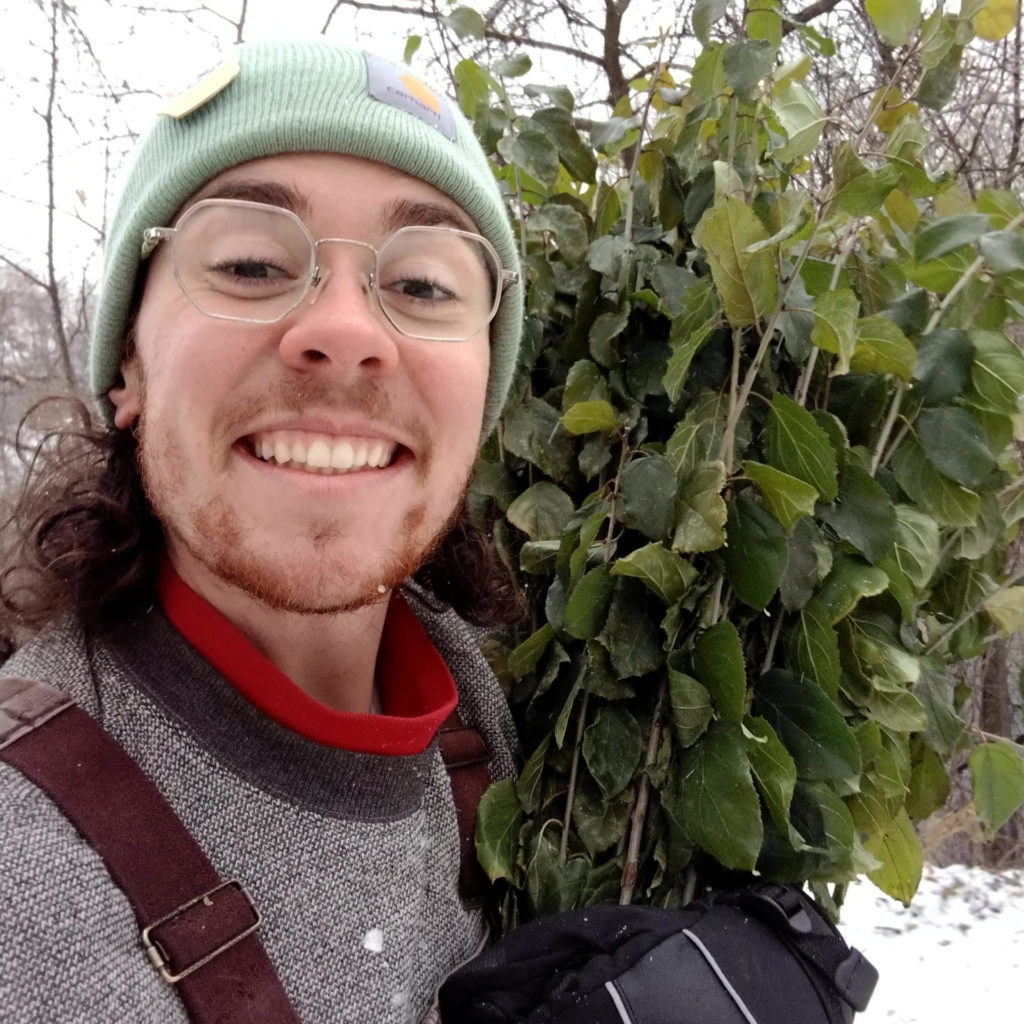 Sashi has glasses facial hair wears wintery clothing and holds greenery against a snowy background