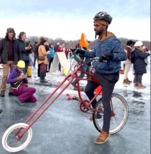 A person in a DIY bike contraption gets ready to race on a frozen lake while onlookers cheer and take photos