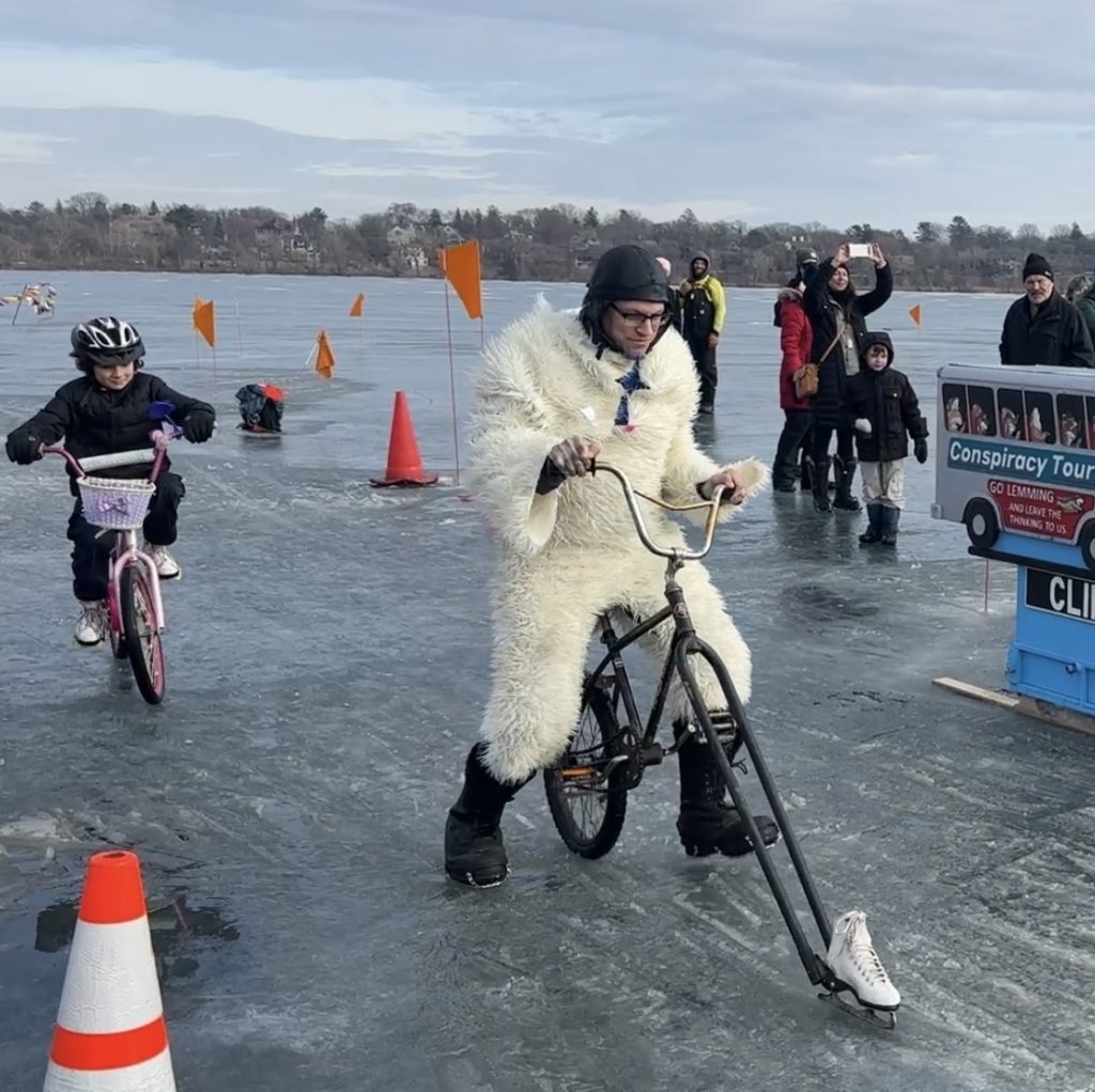 A person in a fluffy white onesie costume leads the pack of frozen lake cyclists while riding a bicycle with an ice skate instead of a front wheel.