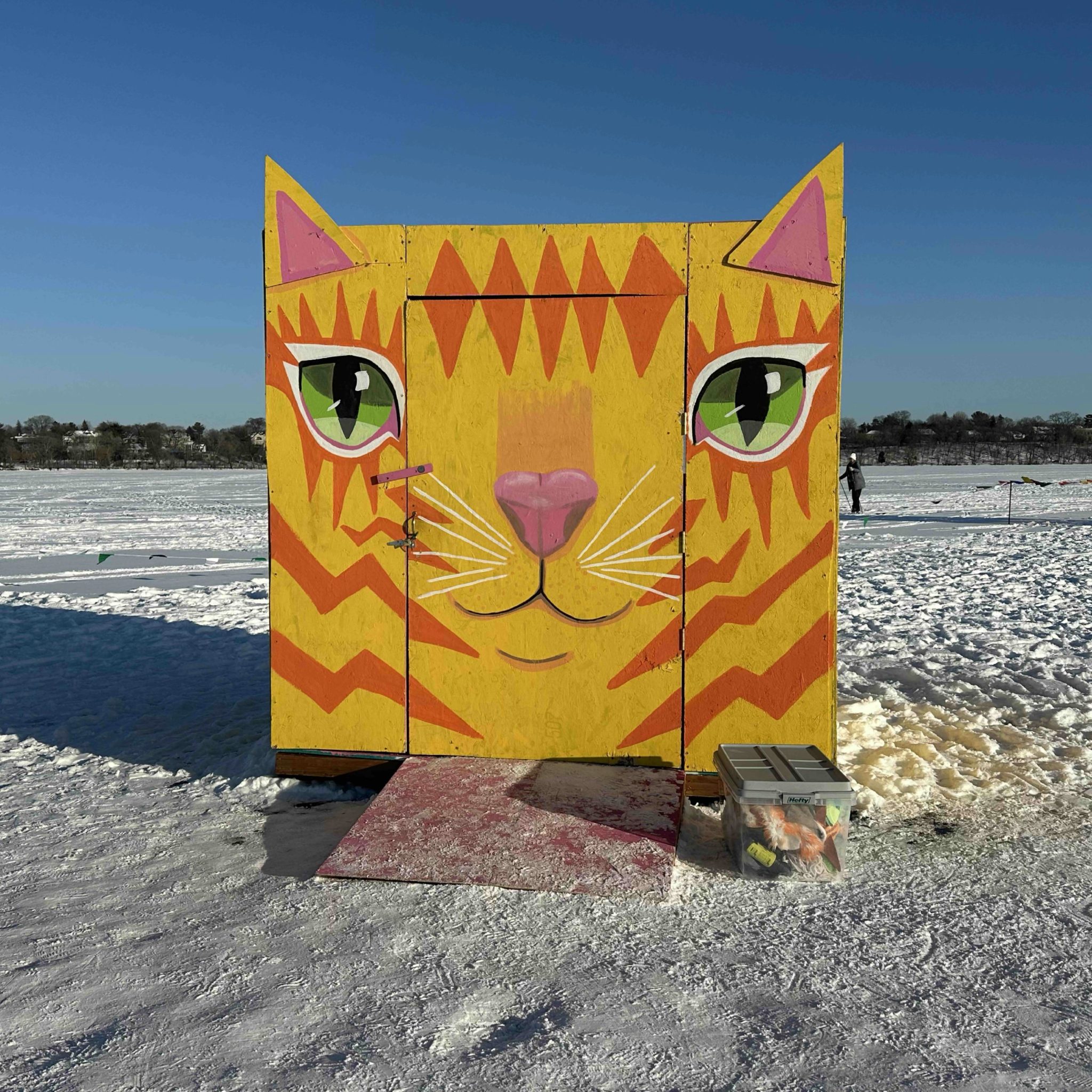 A boxy shanty on a frozen lake painted in bright colors to look like a giant cat face