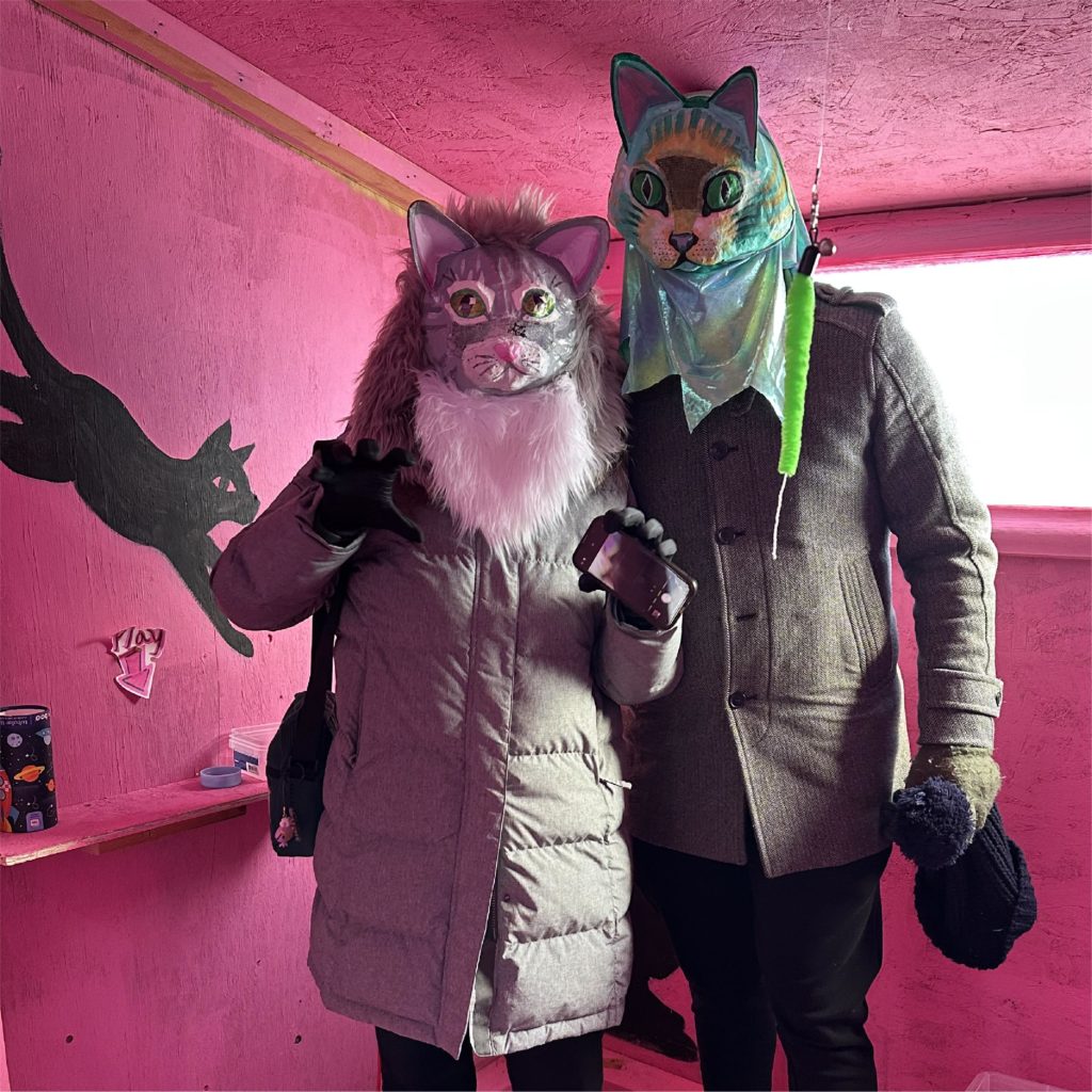 Two people wearing cat masks inside a shanty with pink walls and natural light shining in from behind