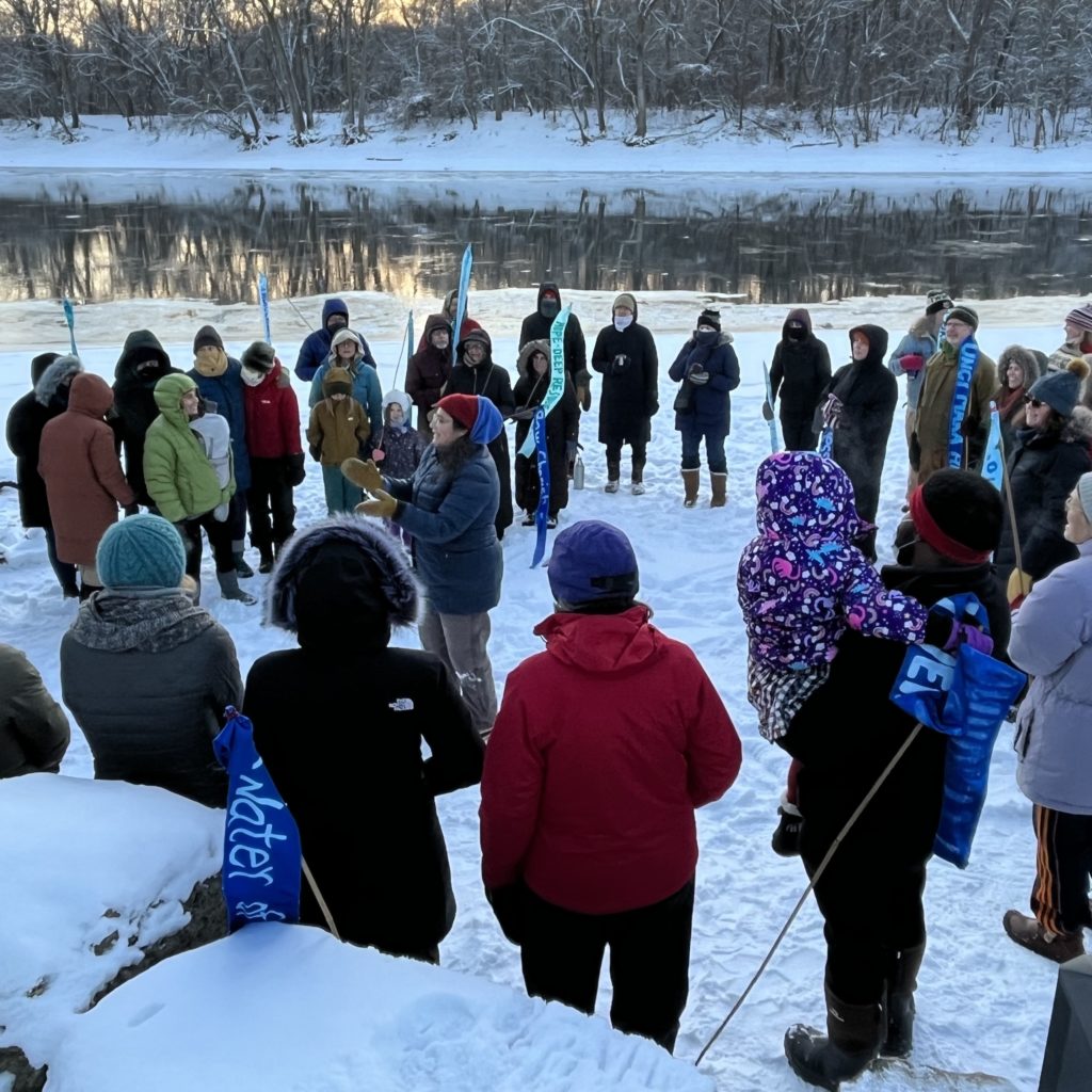 A group of people stand in a circle near a river in wintertime.