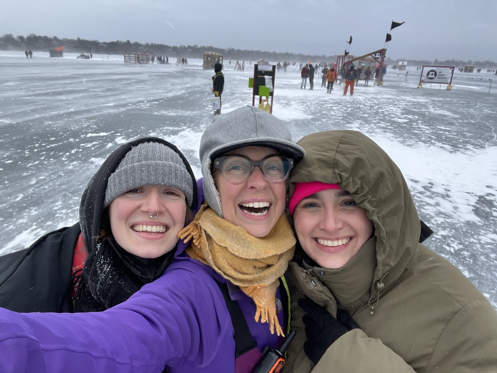 three people take a selfie with big smiles with the shanty village in the distance