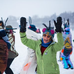 Adults wearing winged costumes and antenna raise their arms in a dance on a frozen lake