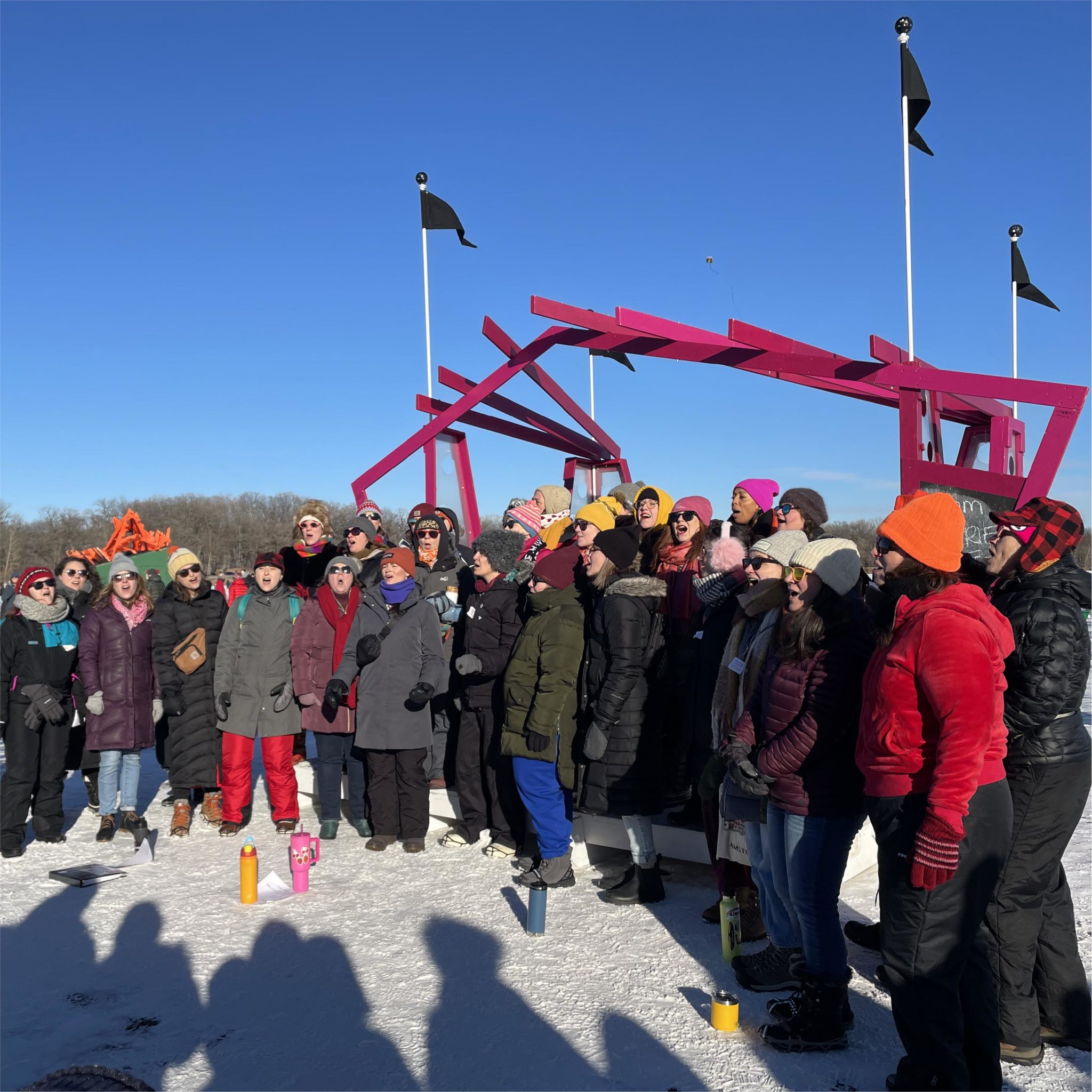 A choir sings outside on a stage on a frozen lake with shadows of the audience across the snowy ground