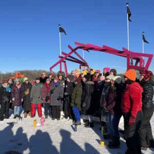 A choir sings outside on a stage on a frozen lake with shadows of the audience across the snowy ground