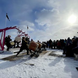 Performers play taiko drums at an outdoor frozen lake stage with a crowd of onlookers in silhouette