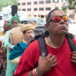 A performer wearing a red shirt and orange tinted sunglasses holds their backpack straps while looking up