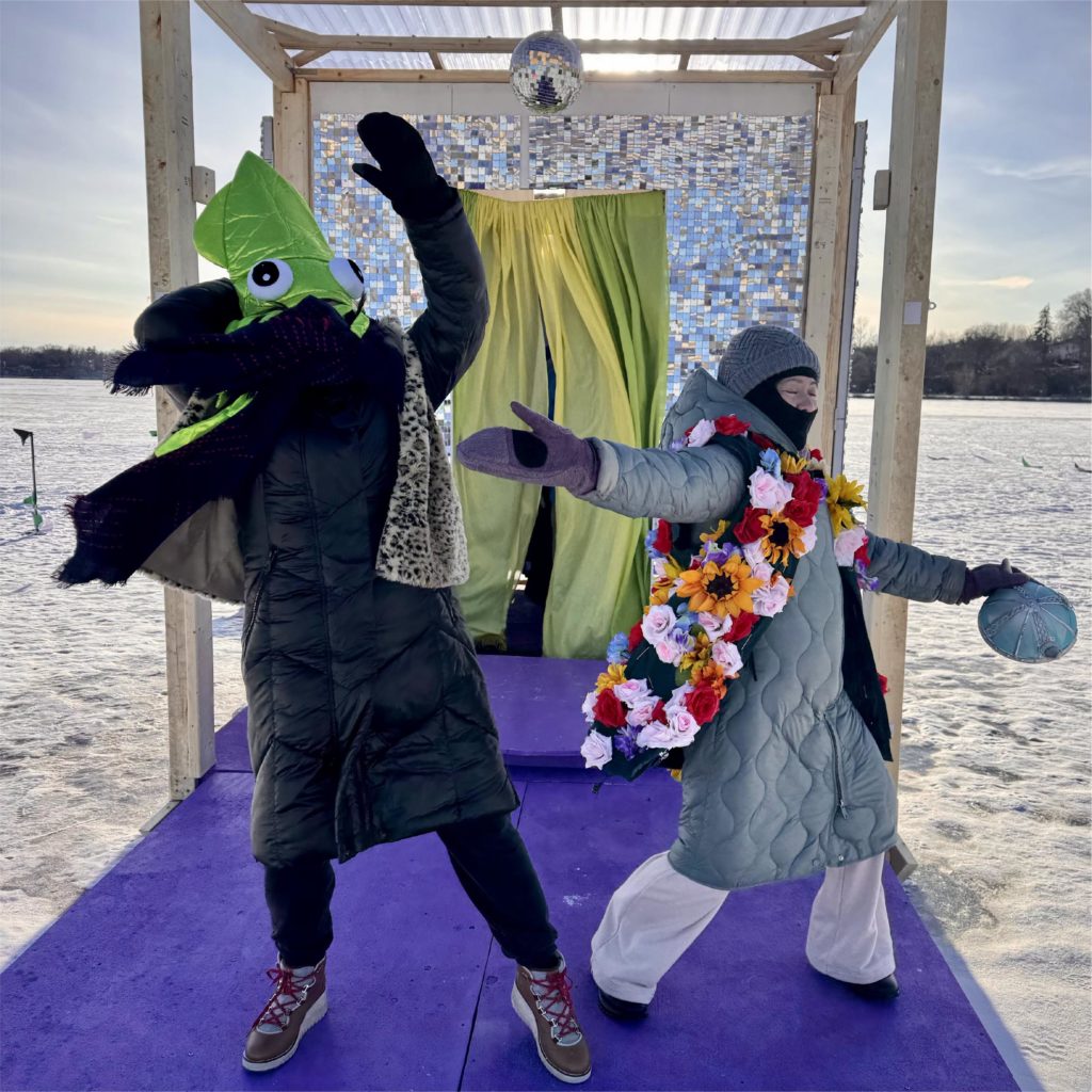 Two people wearing wild costume pieces over their snowsuits, perform on a purple stage underneath a disco ball against a snowy backdrop
