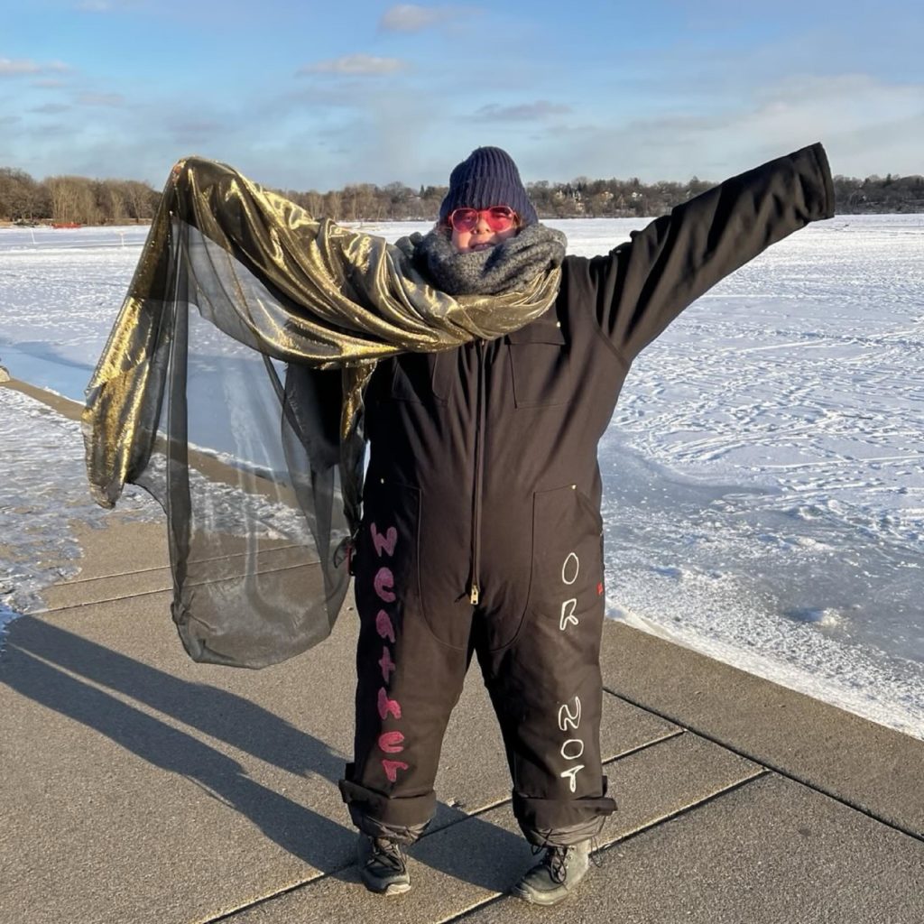 a person in a snowsuit and shimmery gold piece of fabric makes a grand arms out gesture next to the frozen lake