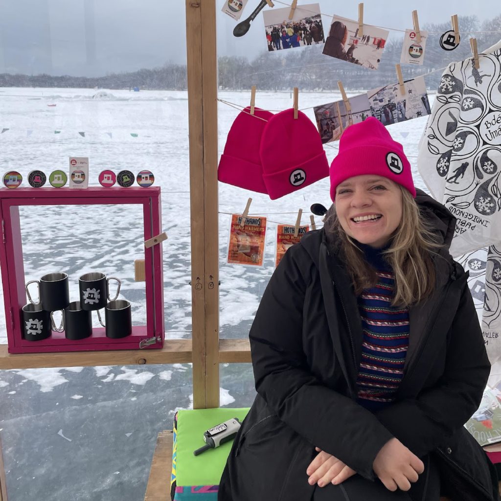 A smiling person wearing a magenta shanty beanie sits inside the Welcome Shanty with the merch display in the background