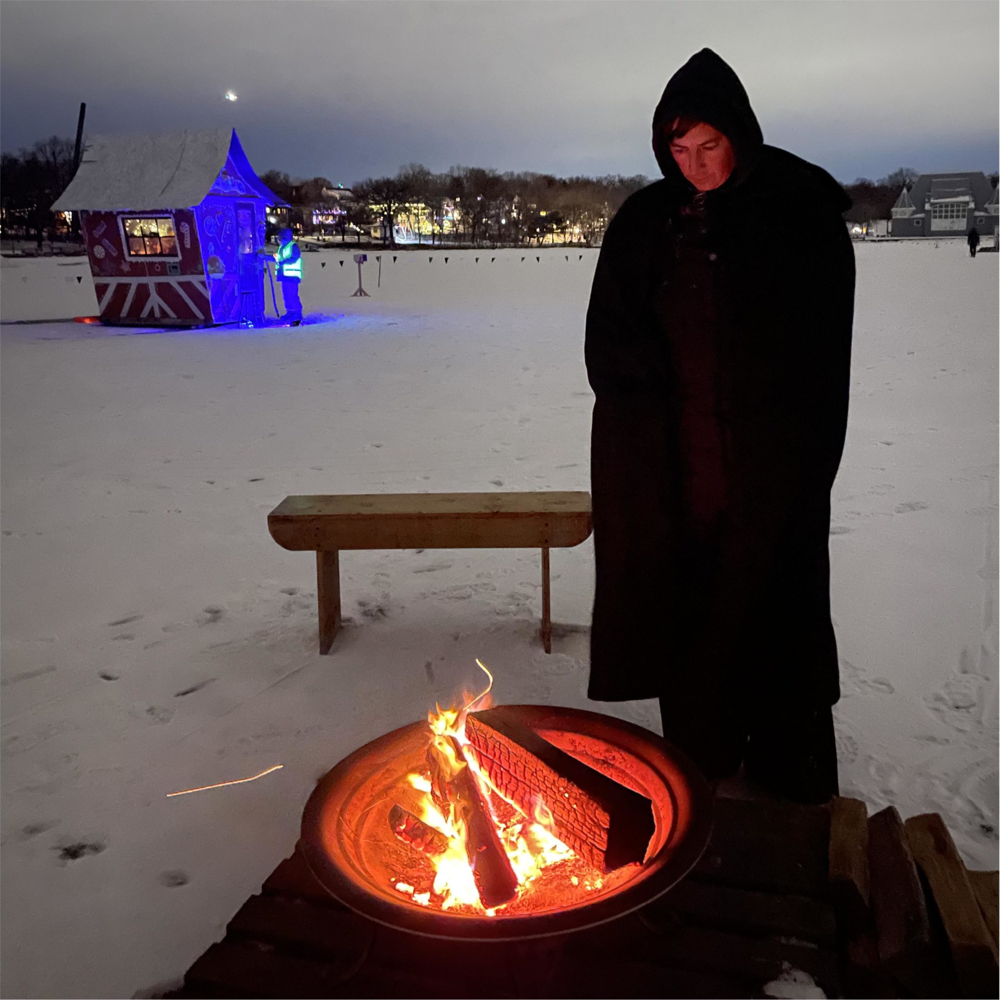 A witch stands in twilight at a raging bonfire. A candy coated gingerbread shanty is in the distance