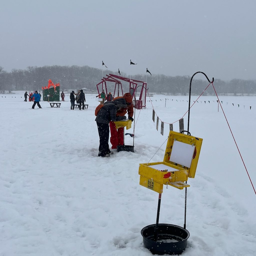 a yellow Berton Box stands with a blank canvas ready to paint on. Plein air painters are dispersed in the background throughout the snowy shanty village