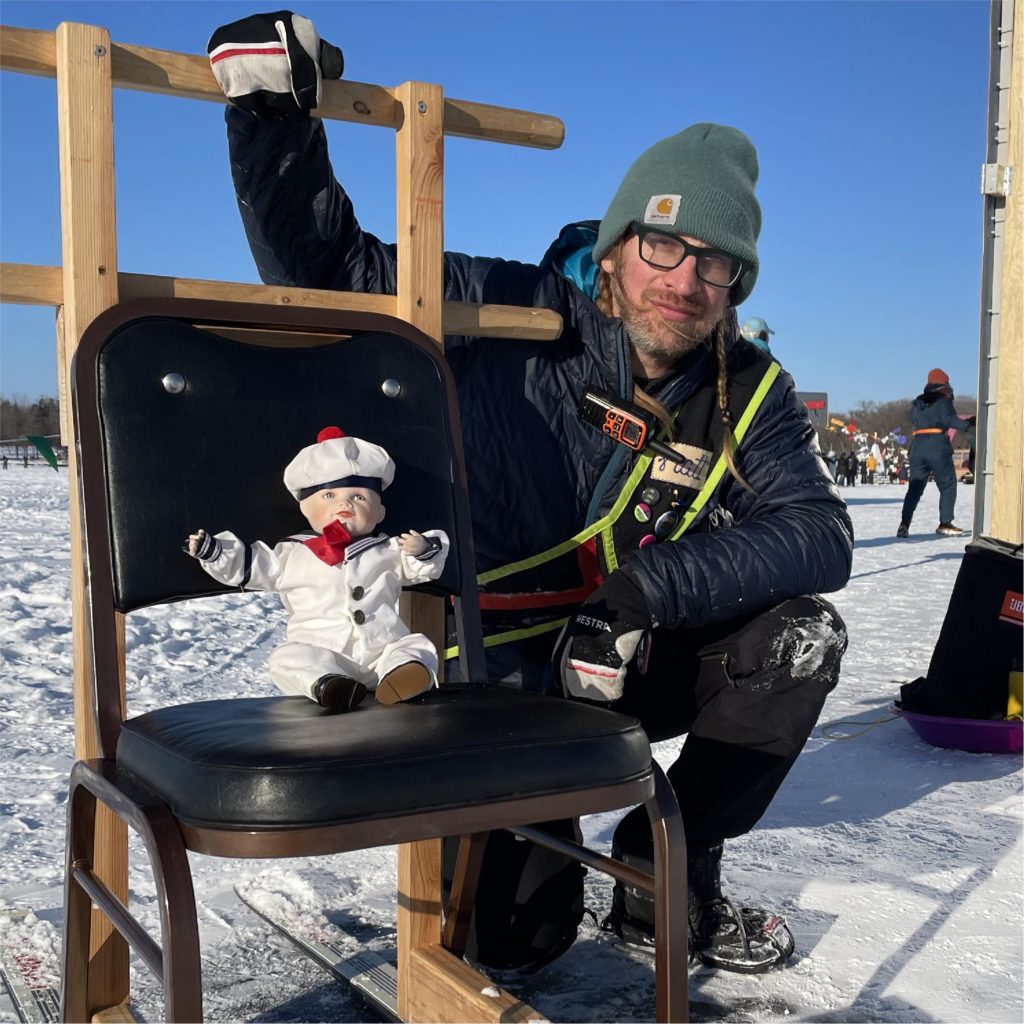 A person wearing a sash kneels next to a kicksled with a sailor doll on the seat