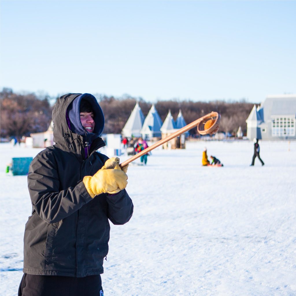 a bunded up person catches a ball in a lacrosse stick on a frozen lake. The blue bandshell on shore is in the far distance