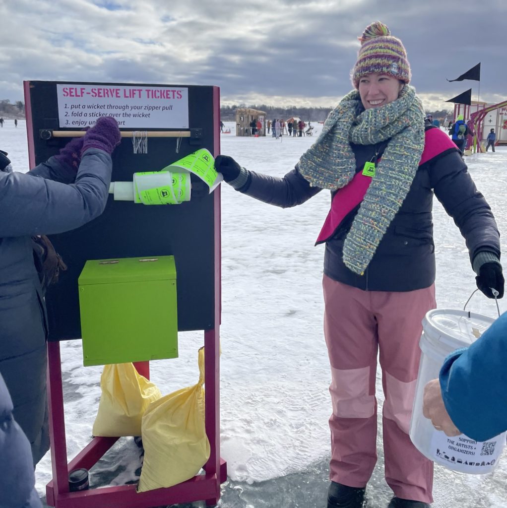 A person in colorful winterwear offers a liift ticket to a shanty visitor