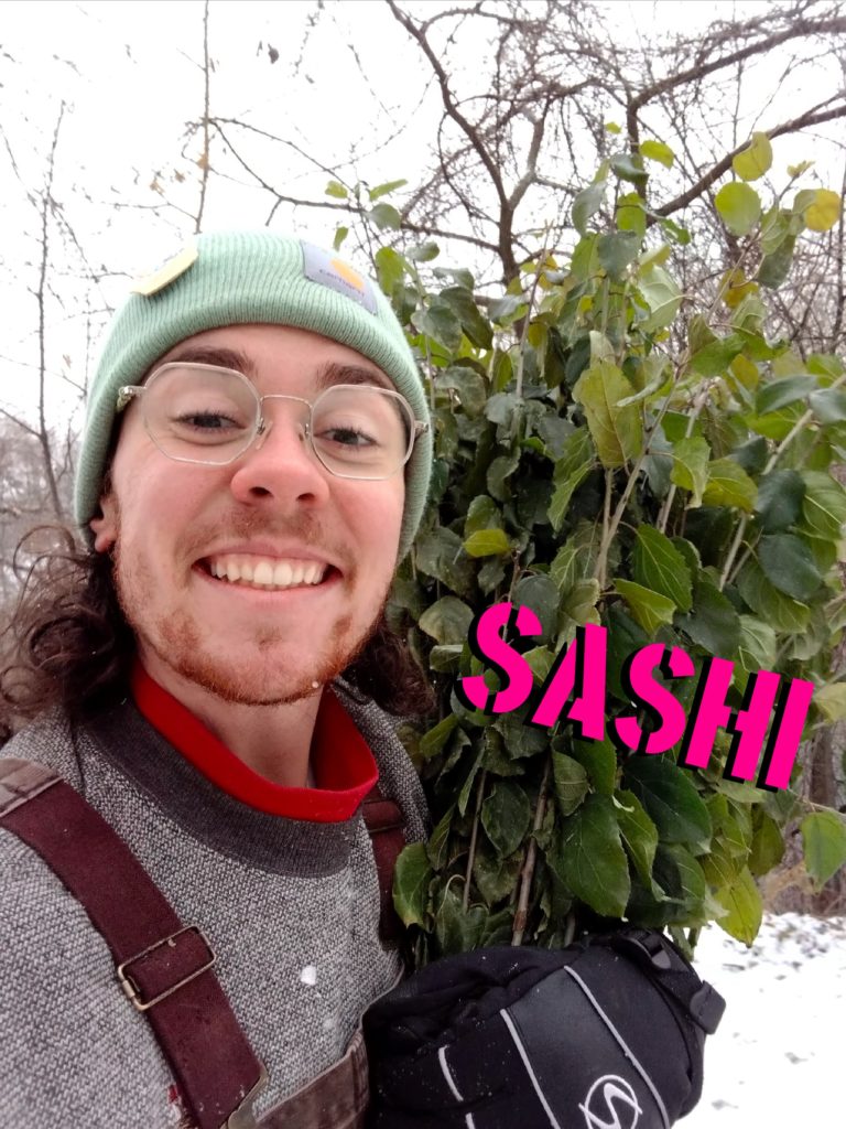 A person with spectacles smiles and holds up winter greenery. Their name, "Sashi" is stenciled across the greenery