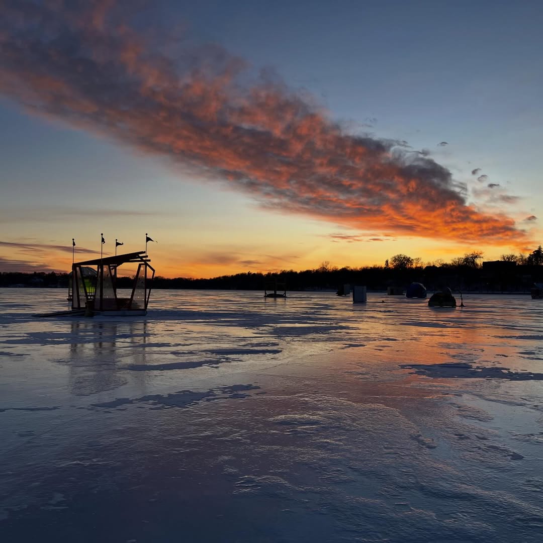 Shanties in silhouette on the frozen lake at sunset. The pinks and oranges of the sky reflect on the shiny lake surface.