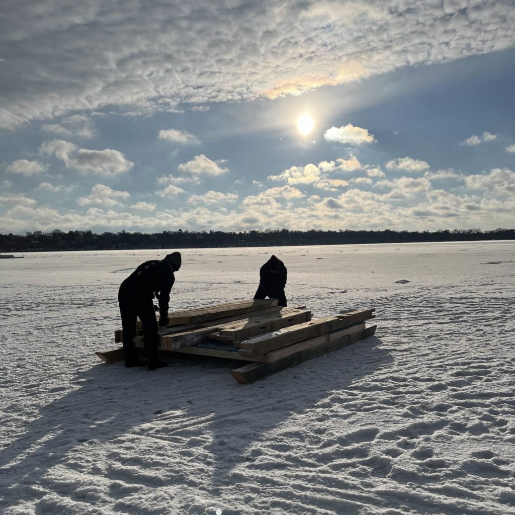 Two people in silhouette add wood to a shanty base in the early morning.