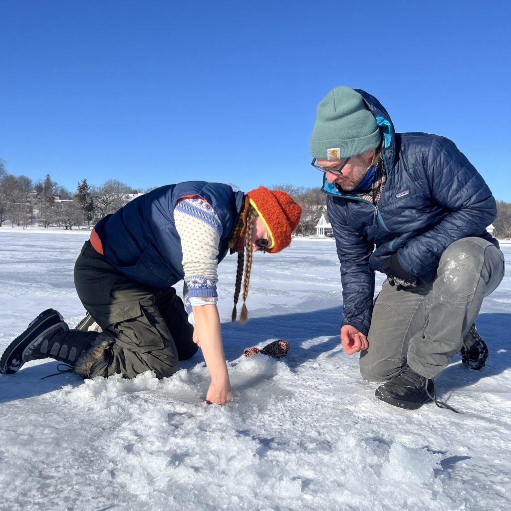 A person with long braids reaches their hand into an auger hole to measure the ice as another person kneels and looks on