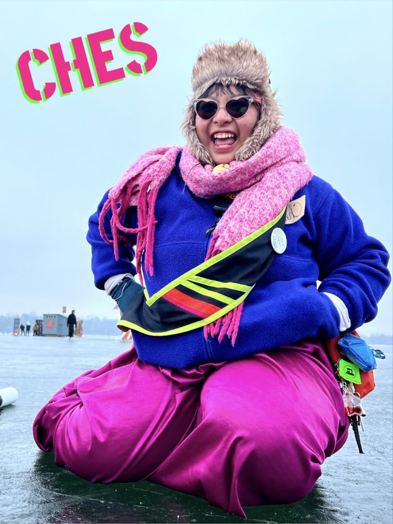 A person wearing brightly colored winterwear and heart shaped sunglasses kneels on the frozen lake. Their name, "ches" is stenciled above